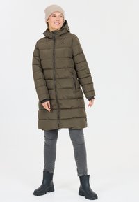 Lungo cappotto piumino verde oliva con chiusura a zip, cappuccio e cuciture orizzontali. Abbinato a jeans grigi e stivaletti neri.