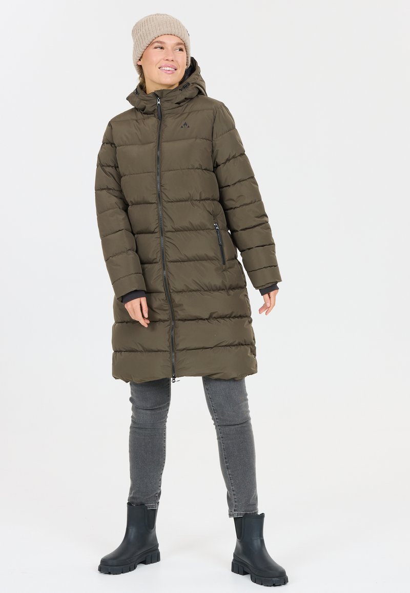 Lungo cappotto piumino verde oliva con chiusura a zip, cappuccio e cuciture orizzontali. Abbinato a jeans grigi e stivaletti neri.