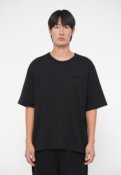 Črna oversize majica iz bombaža, s ribanim krojem in kratkimi rokavi. Diskreten detajl logotipa na sprednji strani. Gladka tekstura.