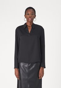 Maglietta nera a maniche lunghe con colletto e scollatura a V, realizzata in un tessuto liscio. Indossata con una gonna in pelle nera aderente.