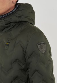 Giacca a vento verde con cappuccio, caratterizzata da un motivo a chevron, dettagli neri e un logo Blauer sulla manica. Tessuto strutturato e trapuntato.