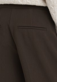 Gros plan sur un pantalon marron sur mesure avec des poches passepoilées à l’arrière et une partie d’un pull en maille crème visible en haut.