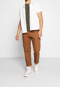 Weißes kurzärmliges Hemd mit Knöpfen, grünes T-Shirt, braune Cargo-Hose und weiße High-Top-Sneaker. Lässiges Ensemble mit gekrempelten Ärmeln.