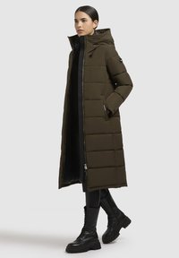 Cappotto imbottito lungo di colore verde oliva con zip anteriore, colletto alto e tasche laterali. Presenta un design trapuntato ed è abbinato a stivaletti neri.