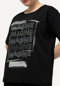 Camiseta de algodón negra con el texto blanco en negrita "IMAGINA" en diferentes estilos y un rectángulo gris que dice "HAY MUCHAS RAZONES PARA SER FELIZ."