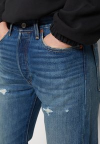 Jeans de mezclilla azul con acabado desgastado, que presentan roturas en los muslos, costuras en contraste y un cierre de botón metálico en la cintura.