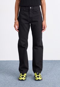 Black denim pants with a straight-leg design, high waist, and front pockets, paired with neon yellow athletic shoes.