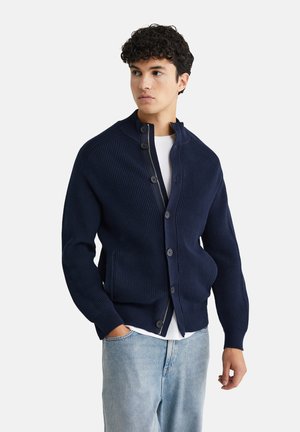 Mornarska rebrasta pletena kardigan s visokim ovratnikom, prednjim gumbima i bočnim džepovima, napravljen od teksturiranog materijala. Nosi se preko bijele košulje.