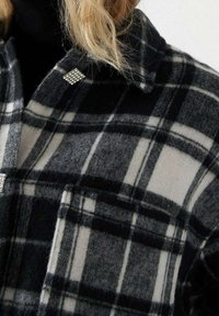 Blond person som bär en svart och vit rutig jacka med rhinestones knappar över en svart polo.