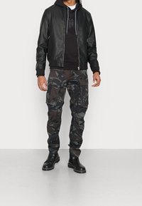 Giacca bomber in pelle nera sopra un sweatshirt con cappuccio nero, abbinata a pantaloni cargo mimetici e stivali alla caviglia in pelle nera.