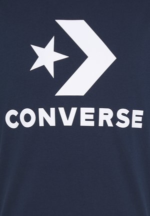 Białe logo Converse z gwiazdą i strzałką nad słowem "CONVERSE" na ciemnoniebieskim tle z tkaniny.