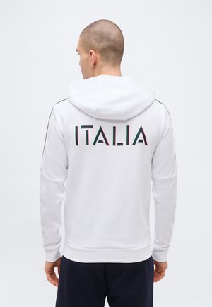 Lühikeste juustega mees, kes kannab valget kapuutsi musta, punase ja rohelise "ITALIA" kirjaga seljal ning musti pükse, näoga eemale.