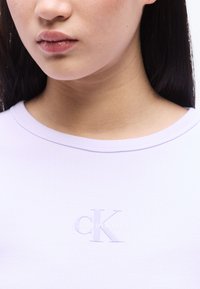 Κοντινό πλάνο στο κάτω μέρος του προσώπου και τον λαιμό μιας γυναίκας που φοράει λιλά ριμπ πλεκτό τοπ με ανάγλυφο λογότυπο Calvin Klein στο στήθος.