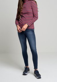 Randig polotröja i röd, vit och marinblå färg, ihopparad med tighta blå jeans och mörka sneakers med vit sula och snörning.