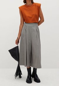 Haut ajusté orange, pantalon large à carreaux, bottines noires, et sac à main noir avec des détails en frange. Les couleurs unies et les motifs contrastent.