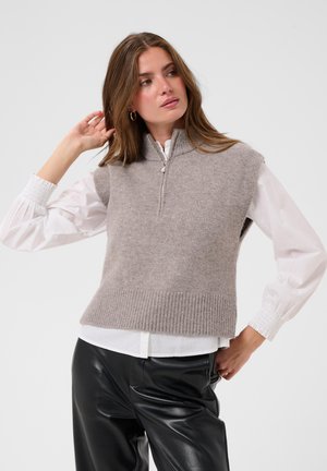 Femme portant un gilet en tricot gris sans manches par-dessus un chemisier blanc à manches longues et un pantalon en cuir noir, avec une main touchant ses cheveux.