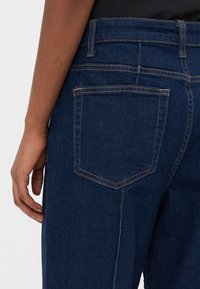 Närbild av mörkblå denimjeans med beige sömmar, med fokus på bakfickan och en persons vänstra hand som vilar bredvid.