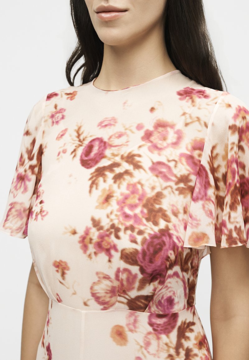Frau trägt ein durchsichtiges, kurzärmeliges Kleid mit rosa und braunen Blumenmustern auf hellbeigem Hintergrund.