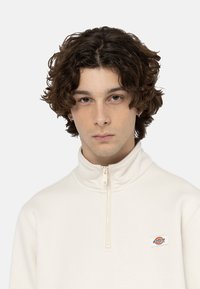 Sudadera blanca con cremallera y alto cuello, de textura suave, con un pequeño parche de logotipo colorido en el pecho, que presenta un diseño minimalista y sencillo.