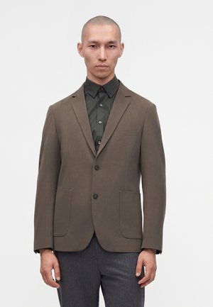 Mand iført en brun blazer over en sort knappet skjorte og grå bukser, stående mod en ensfarvet hvid baggrund.