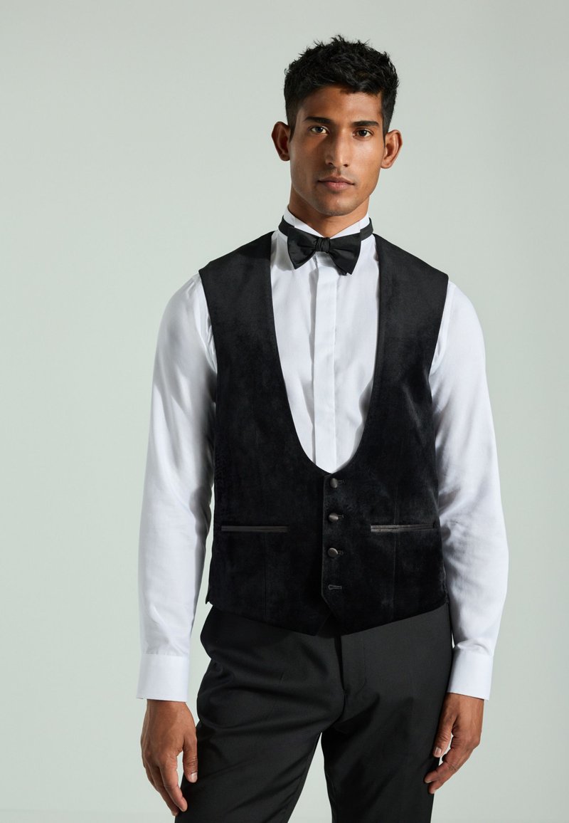 Homme portant une chemise blanche, un nœud papillon noir, un gilet noir boutonné et un pantalon noir, debout devant un fond clair uni.