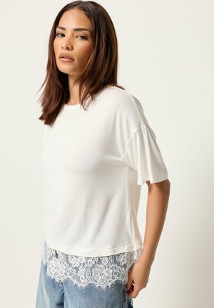 Femme aux cheveux bruns raides vêtue d'un haut blanc à manches courtes avec un ourlet en dentelle associé à un jean bleu, debout devant un fond uni.