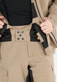 Beige vattentät jacka med dragkedja och tryckknappsstängning, svart foder invändigt och elastisk midja. Fickor synliga på byxorna.