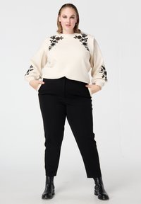 Crèmekleurige sweatshirt met zwarte bloemrijke borduursels, gecombineerd met zwarte taps toelopende broek. Model draagt zwarte enkellaarsjes, handen in de broekzakken.