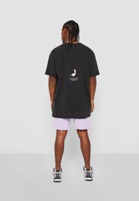 Camiseta negra de gran tamaño hecha de algodón, con un gráfico rosa en la parte posterior, combinada con pantalones cortos violetas claros y zapatillas deportivas negras.