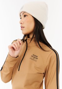 Brązowa bluzka z długim rękawem i kołnierzykiem na zamek błyskawiczny, czarne paski po bokach oraz tekst "HOUSE OF SNOW PROTEST". Połączona z prążkowaną beanie w kremowym kolorze.