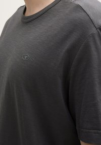 T-shirt gris foncé en tissu doux et texturé. Il présente un col rond et un branding subtil au centre avec "1962" en dessous.