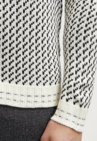 Pull à motif noir et blanc présentant un tricot texturé avec un ourlet et des poignets côtelés. Gros plan sur la section de la manche montrant un design détaillé.