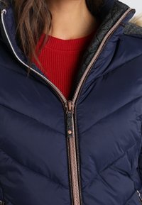 Femme portant une veste matelassée bleu marine avec une fermeture éclair centrale partiellement ouverte, révélant un pull côtelé rouge en dessous.