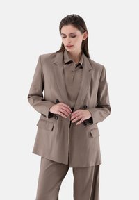Beige Doppelreiher-Blazer mit Revers, vorderen Taschen und schwarzen Knöpfen, kombiniert mit einem passenden Poloshirt in ähnlicher Farbe.