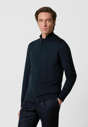 Hombre de piel clara y cabello castaño que lleva un suéter negro con cremallera y pantalones negros, de pie con una mano en el bolsillo contra un fondo blanco.