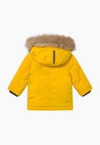 Parka jaune avec capuche bordée de fausse fourrure, accents de poignets bleu marine, étiquette verticale à l'arrière. Caractérisée par une texture lisse et une conception chaude et isolante.