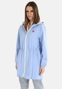 Veste coupe-vent bleu clair avec capuche, taille élastique, fermeture éclair blanche et détail logo. Fabriquée en matériau léger et résistant à l'eau.