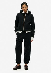 Felpa nera con cerniera e coulisse, abbinata a pantaloni jogger coordinati. L'outfit include una maglietta bianca sotto e mocassini marroni.