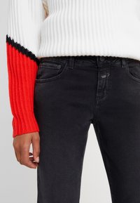 Persona vestida con vaqueros negros y un suéter blanco con mangas a rayas rojas y negras de canalé, con la mano relajada a su lado.