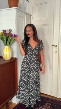 Γυναίκα με μαύρο φόρεμα με λουλούδια και κόκκινο κραγιόν κάνει νόημα προς ένα βάζο με μωβ και κίτρινα λουλούδια σε ένα ξύλινο ντουλάπι μέσα στο δωμάτιο.