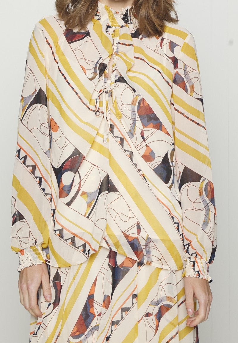 Femme portant une robe à manches longues avec des rayures diagonales jaunes et des motifs géométriques abstraits en tons orange, noir et bleu.