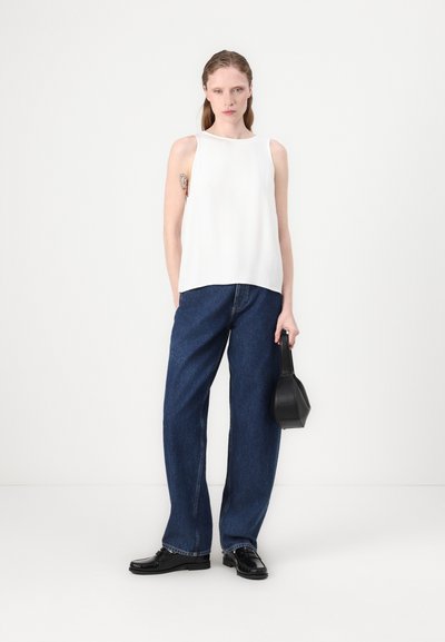 Weißes ärmelloses Oberteil, blaue weiter geschnittene Jeans und schwarze Handtasche. Schwarze Schuhe mit strukturiertem Finish. Sauberes und minimalistisches Design.