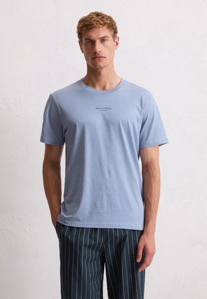 Blonde man in een lichtblauw Marc O'Polo t-shirt en donkere gestreepte broek, staand tegen een effen witte muur.