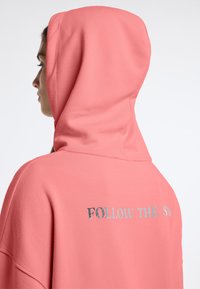 Kapuzenpullover in koraal, met zilveren tekst "VOLG DE ZON" op de achterkant. Gemaakt van zachte stof met een relaxte pasvorm.