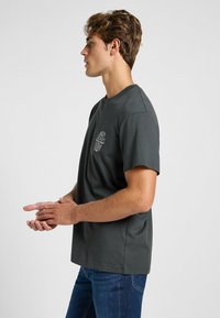 Camiseta de algodón gris oscuro con cuello redondo y mangas cortas, que presenta texto impreso en blanco en el lado izquierdo del pecho. Ajuste casual.