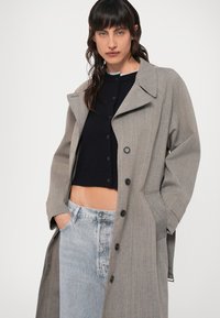 Šedý pruhovaný trenčkot s širokým límcem, velkými černými knoflíky a bočními kapsami, vrstvený přes tmavý cropped svetr s knoflíky a světlé džíny.