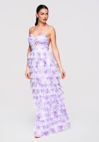 Femme dans une robe à volants superposés lavande et blanche à motifs floraux, avec fines bretelles, posant la tête tournée et les mains détendues sur les côtés.