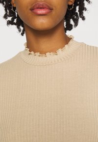Beige gerippter Stricktop mit hohem Rüschenausschnitt. Die Stofftextur ist weich mit einem subtilen Glanz und bietet eine taillierte Silhouette.