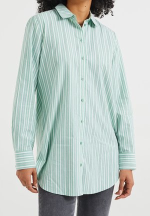 Vrouw die een mintgroen-wit verticaal gestreept overhemd draagt met lange mouwen en grijze broek.
