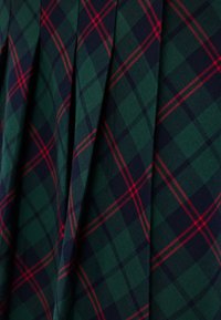 Πράσινο και ναυτικό καρό ύφασμα με κόκκινες λεπτομέρειες, που διαθέτει κομψές πιέτες και υφή. Το μοτίβο αποτελείται από μεγάλα, διασταυρούμενα τετράγωνα.